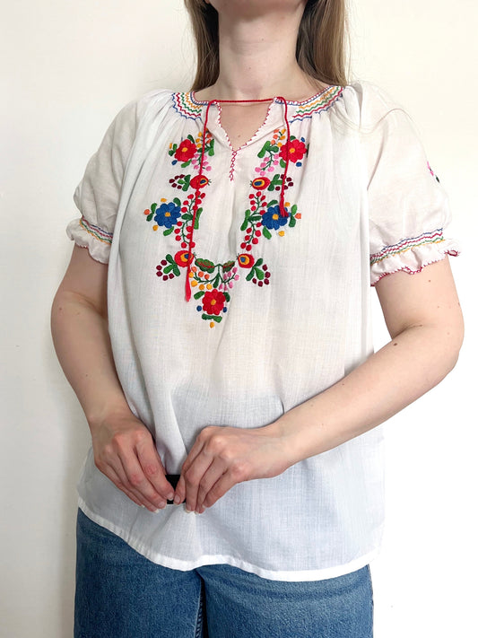 White blouse with colorful embroidery worn by a person on a plain background